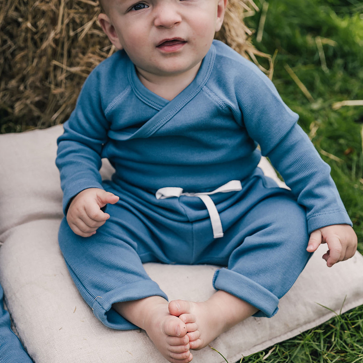 baby sitting on pillow on grass wearing uaua kimono onesie and joggers in azul color