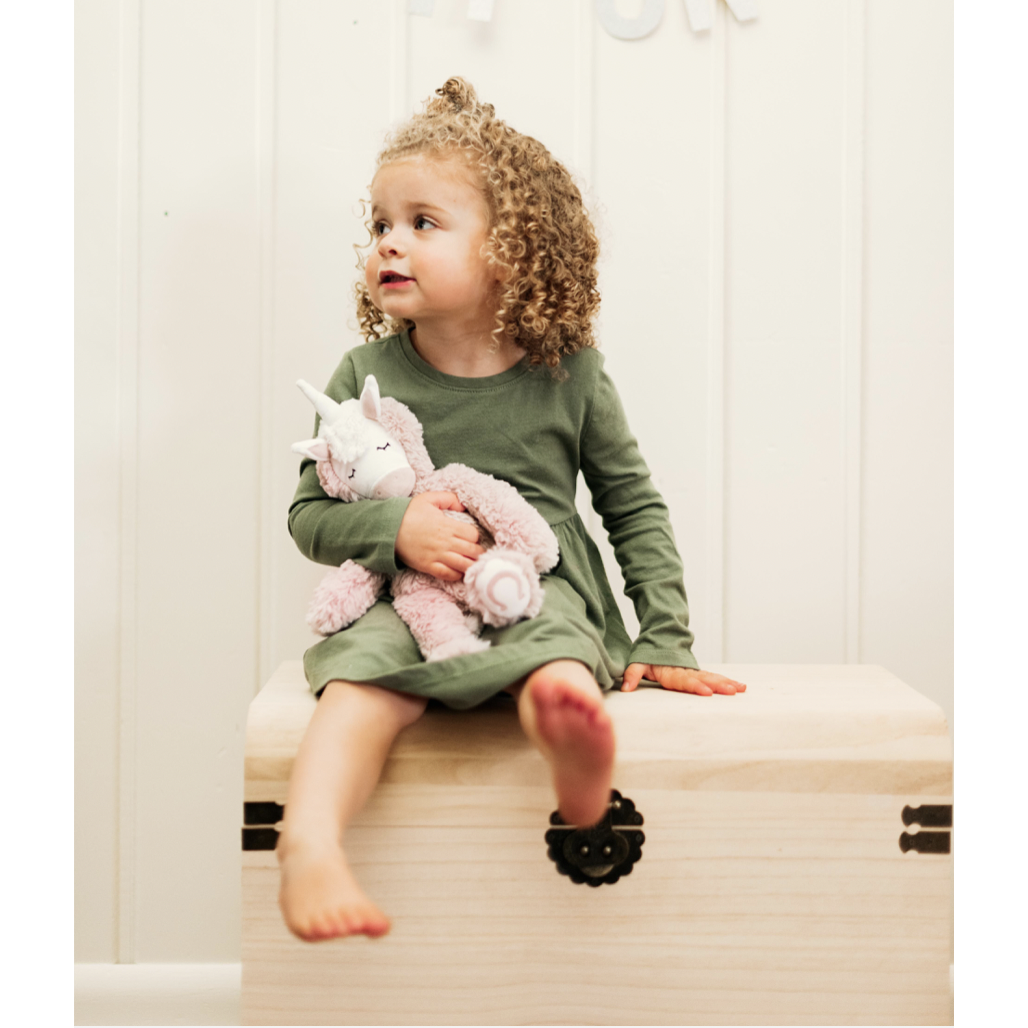 curly haired little girl in green dress sitting on bench holding slumberkins pink unicorn kin