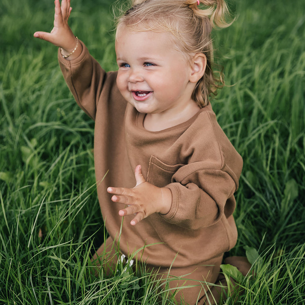 little girl kneeling in grass waving, wearing uaua sweater in chocolate