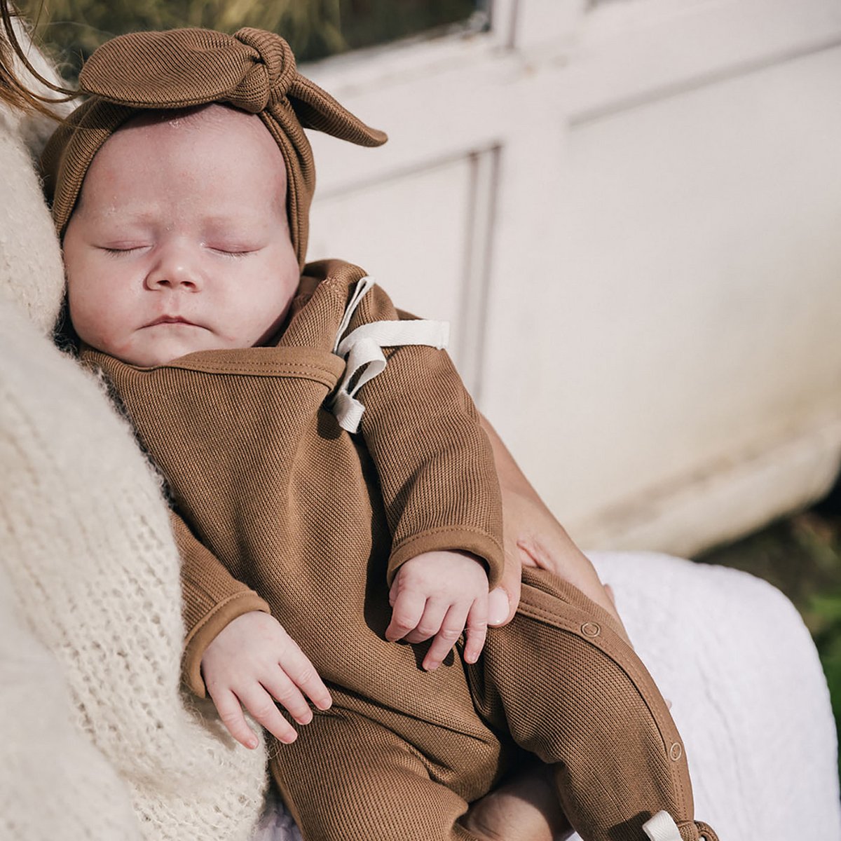 Sleeping baby in mothers arms wearing uaua kimono style footed romper with bow in chocolate with matching knotted headband